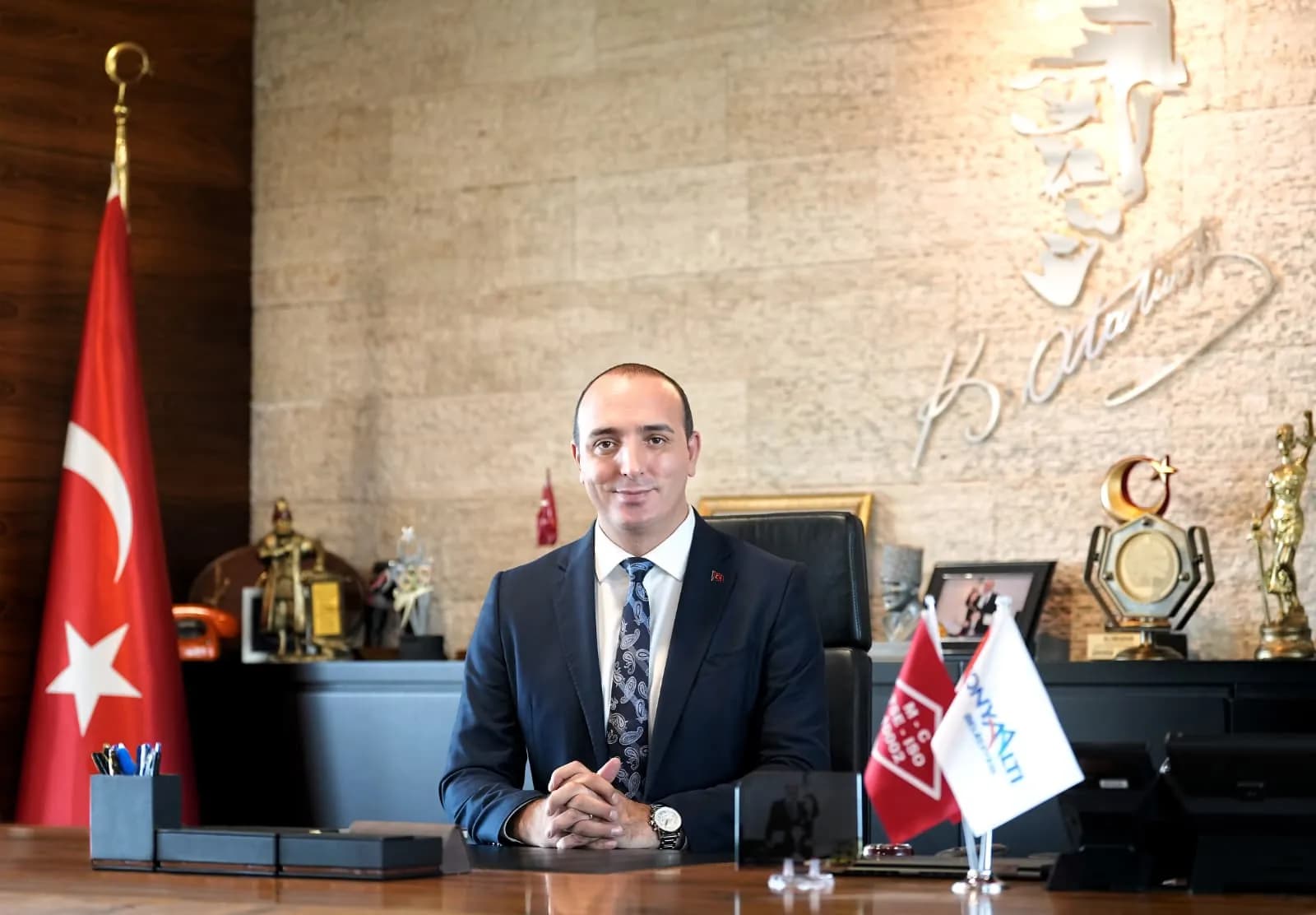 Konyaaltı Belediye Başkanı Cem Kotan ellerini birbine bağlamış takım elbiseli gülümseyerek belediye makamında oturuyor