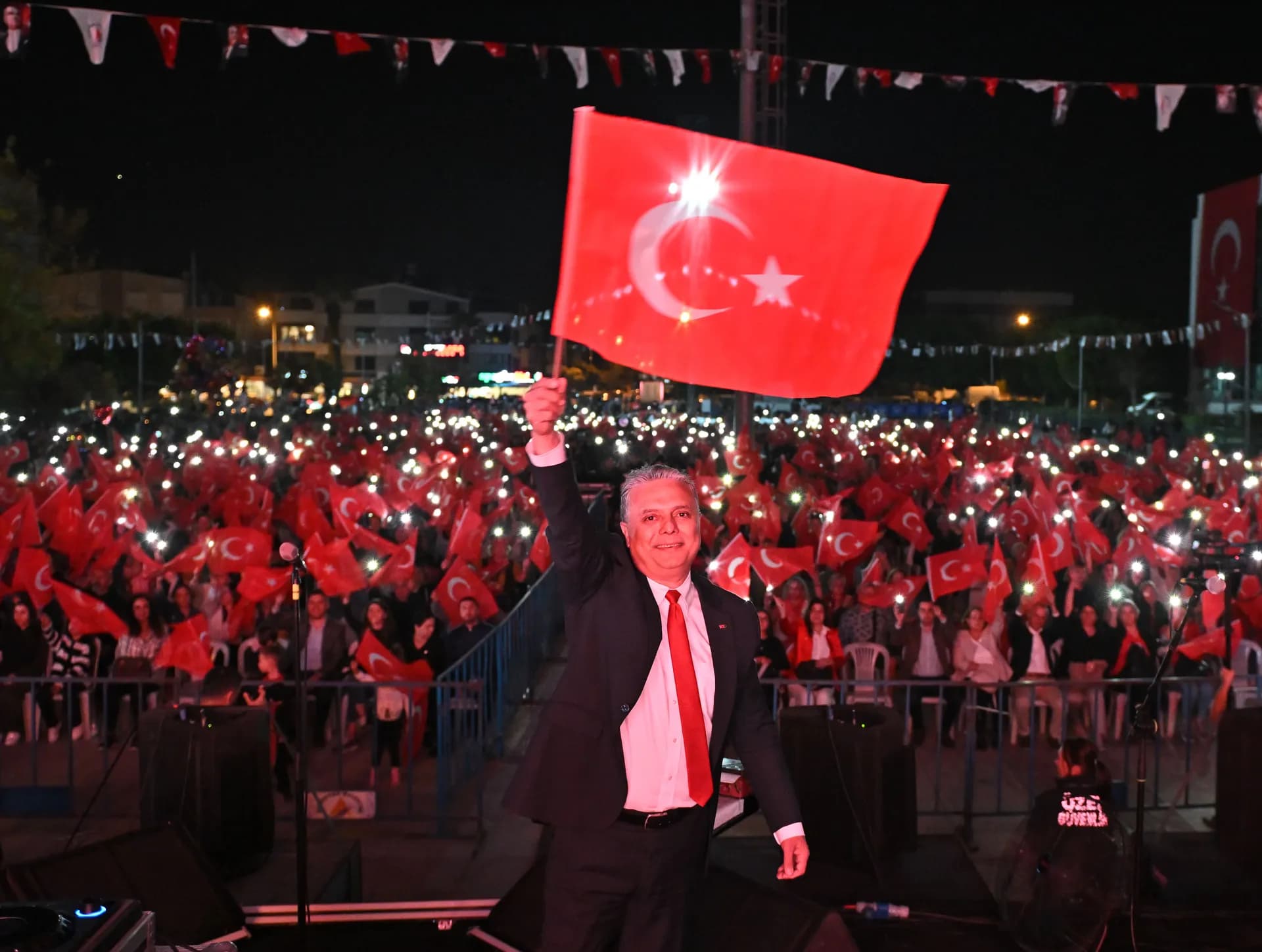 Muratpaşa Belediye Başkanı Uysal, elinde Türk bayrağı tutuyor. Uysal'ın üstünde siyah takım elbise kırmızı kravat arkasında ellerinde bayrak olan kalabalık topluluk yer alıyor