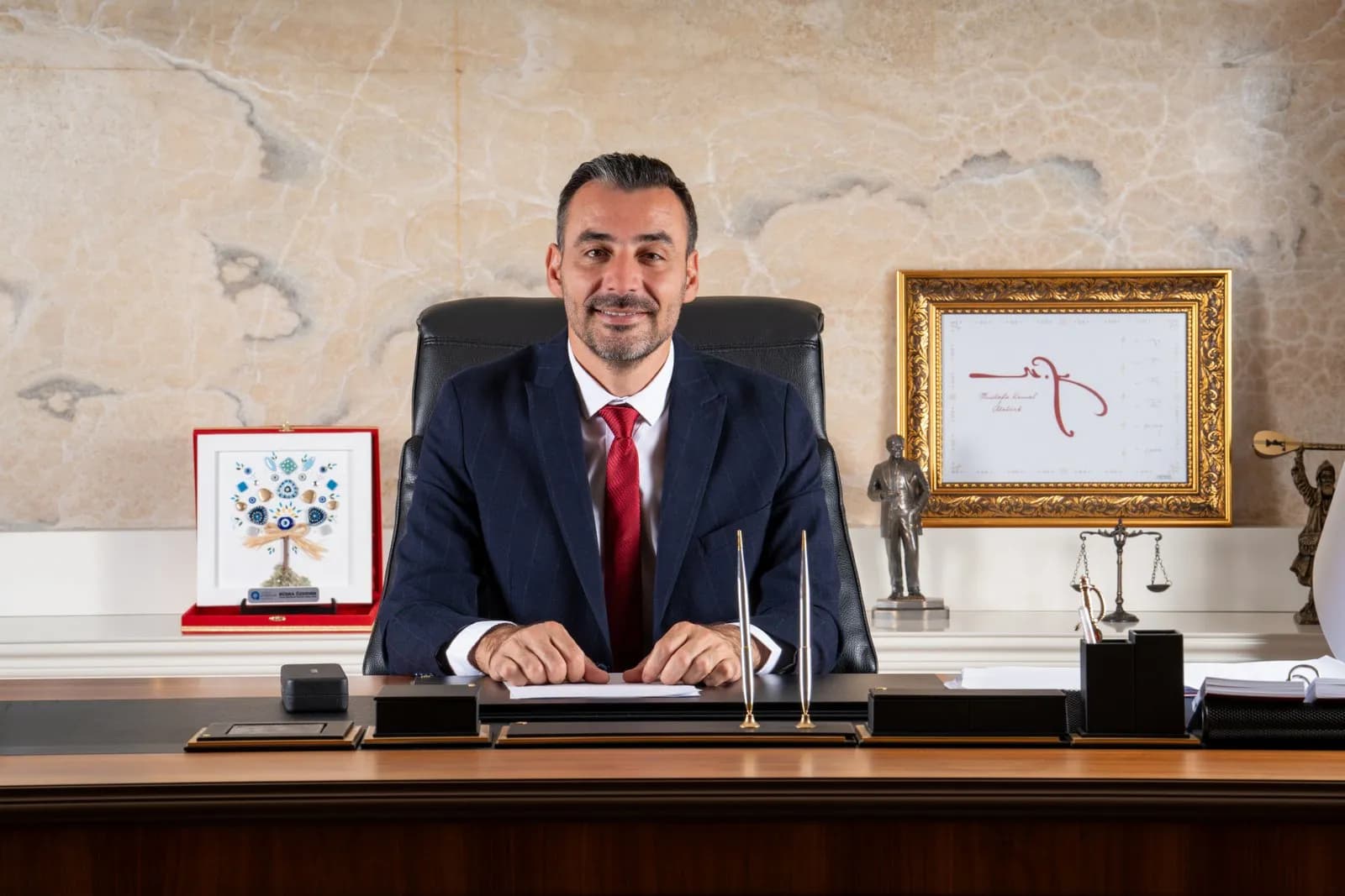 Manavgat Belediye Başkan Vekili Çiçek takım elbiseli otururken gülümsüyor elleri önünde kırmızı kravatı var