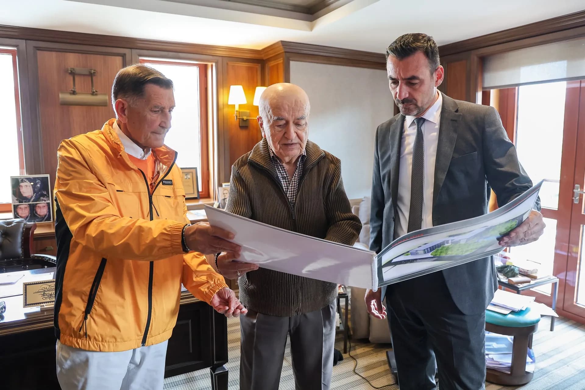 Üç erkek ellerinde büyük proje dosyasına bakıyor. Sağdaki erkek takım elbiseli orta yaşlarda, ortadaki yaşlı beyaz saçlı diğeri de orta yaşlarda 