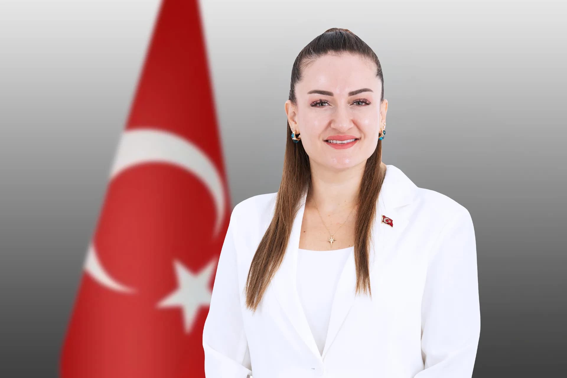 Başkan Vekili Büşra Özdemir beyaz ceket içinde beyaz bluz yanında Türk bayrağı ayakta gülümseyerek duruyor